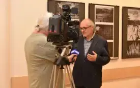 Mężczyzna w okularach i ciemnej marynarce udziela wywiadu przed kamerą stacji TVP3, stojąc na tle czarno-białych fotografii w Galerii Pod Kopułą.