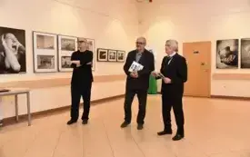 Trzej mężczyźni stoją w przestrzeni wystawienniczej i przemawiają podczas wernisażu wystawy fotografii, kt&oacute;rej prace wiszą na ścianach w tle