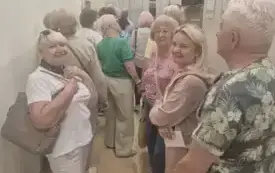 Pomieszczenie Magazynu Książek. W wąskim korytarzu stoją słuchacze Uniwersytetu Trzeciego Wieku. Po prawej stronie widać wysokie szare regały z książkami.
