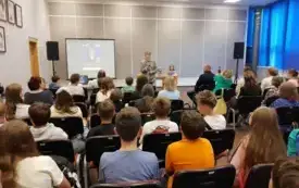Publiczność zgromadzona w sali. Z tyłu rzutnik z plakatem wydarzenia, autorka oraz prowadząca