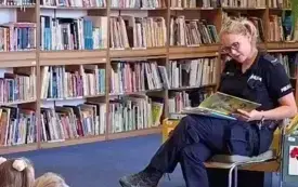 Policjantka siedzi na krześle i czyta, za nią znajdują się regały z książkami. Na starym katalogu bibliotecznym stoi Plastuś &ndash; patron naszej biblioteki. Dzieci siedzą naprzeciwko gościa na dywanie i uważnie słuchają. 
