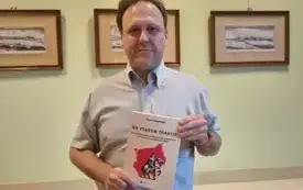 Na zdjęciu autor stoi na tle zielonej ściany sali, na kt&oacute;rej powieszone są dawne ryciny przedstawiające miasta z regionu. Autor prezentuje swoją książkę trzymając ją przed torsem. Patrzy w obiektyw aparatu i się uśmiecha.