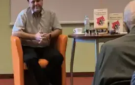 Zbliżenie na autora, zdjęcie zrobione od frontu. Autor patrzy w kierunku aparatu i się uśmiecha. Obok niego stolik