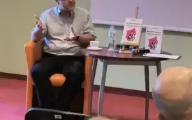 Zbliżenie na autora, zdjęcie zrobione spod prawej ściany sali, lekko pod kątem. Autor siedzi w fotelu i m&oacute;wi gestykulując, wzrok kieruje w prawą stronę zdjęcia, na moderatora. 