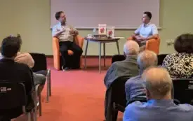 Na pierwszym planie zdjęcia widać uczestnik&oacute;w spotkania.  Na drugim planie, w centralnej części zdjęcia znajdują się (od lewej patrząc) autor książki, stolik z wyeksponowanymi dwoma egzemplarzami książki oraz moderator spotkania.