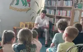 Na jasnej ścianie emblemat biblioteki, regały z kolorowymi książkami, przed tym siedzący mężczyzna czytający książkę. Przed nim grupa zwr&oacute;conych w jego kierunku dzieci. Na bokach regał&oacute;w z książkami plakaty. W tle zielone rośliny.