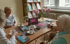 Siedzące przy stole panie, na brązowym obrusie książki, fioletowe emblematy DKK, filiżanki z napojami, słodycze , wazon z r&oacute;żowymi kwiatami. W tle regał z książkami.