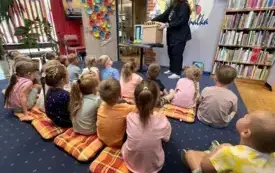 Kobieta pokazuje rekwizyty grupie dzieci siedzącej na dywanie