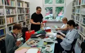 Na zdjęciu widać trzy uczestniczki zajęć siedzące przy stole, kt&oacute;re wykonują swoją pracę i prowadzącą zajęcia, Paulinę Wesołowską, kt&oacute;ra stoi i pokazuje jak coś wyciąć. Na stole są białe filiżanki, talerzyki, materiały plastyczne: ozdobne papiery, noży