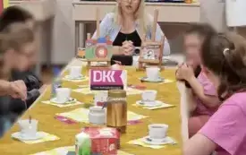Na ścianie kolorowe plansze, w tle st&oacute;ł z dekoracjami, a przy stole siedząca osoba.