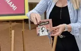 Osoba trzyma w dłoni audiobook "Album z emocjami", w tle sztaluga z plakatem DKK oraz st&oacute;ł.