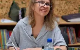 Pisarka siedzi przy stole. Uśmiecha się. W prawej ręce trzyma długopis. Za nią wiszą kolorowe prace.