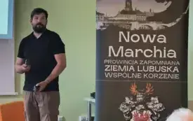 Zbliżenie na prelegnta stojącego przy rollupie z pełną nazwą cyklu spotkań &bdquo;Nowa Marchia &ndash; Prowincja zapomniana &ndash; Ziemia Lubuska &ndash; Wsp&oacute;lne korzenie&rdquo;. Prelegnt jest zwr&oacute;cony do kamery lekko bokiem, wzrok kieruje w prawo, na uczestnik&oacute;w spotkania.
