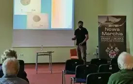 Na pierwszym planie widać uczestnik&oacute;w spotkania siedzących na krzesełkach. W tle stoi prelegent i opowiada patrząc w stronę słuchaczy. Na wyświetlonym slajdzie prezentacji widać zdjęcie monet. Po prawej stronie znajduje się rollup z pełną nazwą cyklu spot