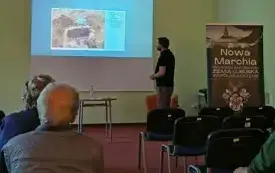 Zdjęcie wykonane jest tak samo jak poprzednie. R&oacute;żni się slajd prezentacji, teraz przedstawione są zdjęcia monet. Prelegent omawia je patrząc w stronę słuchaczy.