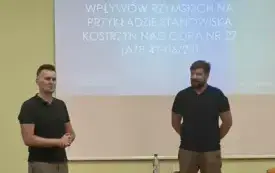 Kierownik Działu Zbior&oacute;w Regionalnych, rozpoczynający spotkanie oraz prelegent. Obaj mężczyźni stoją. Między nimi znajduje się stolik, na kt&oacute;rym są butelka z wodą, szklanka i pilot do obsługi rzutnika. Na ścianie za nimi jest wyświetlona niebieski slajd t