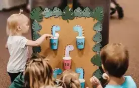 Dzieci biorą udział w zabawie &bdquo;koszyk&oacute;wka z dinozaurami&rdquo;, wrzucając piłki do koszyczk&oacute;w. 