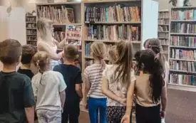 Bibliotekarka prezentuje przedszkolakom wybrane książki znajdujące się na bibliotecznych regałach.