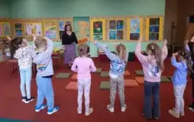 Dzieci stoją. Mają podniesione ręce. Przed nimi stoi bibliotekarka. Za nią na ścianie wiszą kolorowe prace.