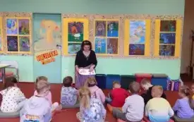 Zer&oacute;wkowicze siedzą na poduszkach. Przed nimi siedzi bibliotekarka. Pokazuje ilustracje w książce. Za nią na ścianie wiszą kolorowe prace.