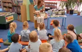 Dzieci siedzą na dywanie przed bibliotekarką, kt&oacute;ra czyta im bajkę o smerfach oraz pokazuje obrazek z książki. Obok niej stoi stoliczek z książkami o smerfach.