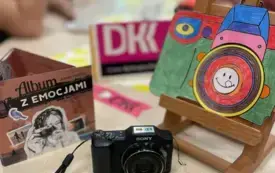 Okładka audiobooka pt.: Album z emocjami Ariadny Piepi&oacute;rki, czarny aparat fotograficzny, kolorowa malowanka aparat fotograficzny. W tle skr&oacute;t DKK i młodzież siedząca przy długim stole.
