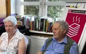 Dwie osoby siedzą przy stoliku, w tle okno oraz plakat z logiem DKK.