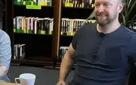 Osoba siedzi przy stoliku, w tle regał na książki.