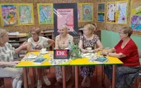 Pięć czytelniczek siedzi przy stole. Rozmawiają o książce. Za nimi jest sztaluga z plakatem DKK i kolorowe prace plastyczne.
