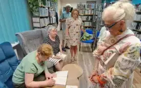 Jedna osoba siedzi i podpisuje książkę, a druga stoi naprzeciwko. 