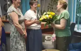 Trzy osoby stoją obok siebie, jedna z nich wręcza kwiaty.