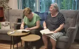 Dwie osoby siedzą przy stoliku, w tle duże okno wraz z roletami.