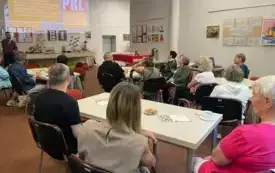 Osoby siedzące przy stolikach, w tle prezentacja multimedialna, przy kt&oacute;rej stoi osoba z mikrofonem