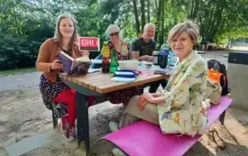Czw&oacute;rka klubowicz&oacute;w Dyskusyjnego Klubu Książki siedzi przy drewnianym stoliku na plaży (trzy kobiety, jeden mężczyzna). Jedna z uczestniczek trzyma otwartą książkę &bdquo;Zgiełk serca. Opowieść o Marii Pawlikowskiej-Jasnorzewskiej&rdquo; Małgorzaty Czyńskiej oraz 