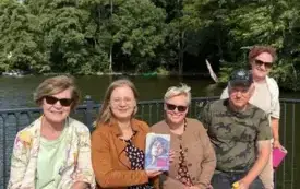 Piątka uczestnik&oacute;w Dyskusyjnego Klubu Książki siedzi na ławce nad jeziorem w Lubniewicach. Uśmiechają się . Jedna z uczestniczek trzyma w g&oacute;rze książkę &bdquo;Zgiełk serca. Opowieść o Marii Pawlikowskiej-Jasnorzewskiej&rdquo; Małgorzaty Czyńskiej. Za klubowiczami 