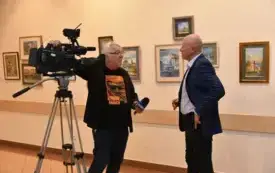 Operator telewizyjny przeprowadza wywiad z akwarelistą Klaudiuszem Pohlem na tle prezentowanych na wystawie obraz&oacute;w przedstawiających Gorz&oacute;w Wielkopolski.