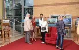 Agata Kaczuk-Jagielnik podczas wernisażu wystawy &bdquo;Miasto w kadrach fotografa i malarki&rdquo; wskazuje prezentowane prace. W tle widoczne są jej obrazy olejne przedstawiające panoramy Gorzowa Wielkopolskiego oraz przestrzeń łącznika Biblioteki Herberta