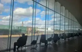 Osoby siedzące na krzesłach, w dłoni trzymający książki, wszyscy patrzą przez szybę.