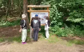 Na tle zielonej roślinności i pni drzew tablica informacyjna przed nią grupka dzieci