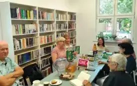 Przy stole siedzi siedmioro uczestnik&oacute;w Dyskusyjnego Klubu Książki: cztery kobiety i trzech mężczyzn. Dwie kobiety po lewej stronie trzymają książki Aurory Tamigio w g&oacute;rze. Jeden z czytelnik&oacute;w po prawej stronie czyta książkę