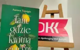 Na białej p&oacute;łce stoi książka Aurory Tamigio &bdquo;Tam, gdzie kwitną cytryny&rdquo;. Obok niej, po prawej stronie stoi mała sztaluga. Na niej jest logo Dyskusyjnego Klubu Książki. Widać kawałek kolejnej białej p&oacute;łki, kt&oacute;ra wisi nad książką. 