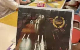 Na pierwszym planie opakowanie od audiobooka, w tle długi st&oacute;ł i siedzące osoby dorosłe.