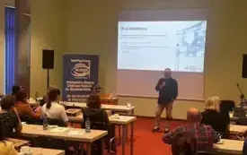 Osoby siedzące w ławkach, w pomieszczeniu panuje jasność.