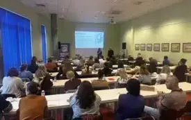 Osoby siedzące w ławkach, w pomieszczeniu panuje jasność.