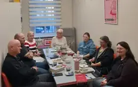 Kameralne spotkanie w pokoju bibliotecznym. Przy długim stole siedzi siedem os&oacute;b. Uczestnicy są zwr&oacute;ceni ku sobie, rozmawiają i uśmiechają się. Na stole widoczne są filiżanki, dzbanek, talerzyki z poczęstunkiem oraz kilka egzemplarzy książki. 
