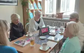 Siedzące przy stole panie, na brązowej serwecie książki, fioletowe emblematy DKK, filiżanki z napojami, słodycze. W tle na parapecie zielone kwiaty w doniczkach, regał z książkami.