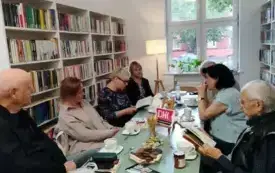 Si&oacute;demka uczestnik&oacute;w Dyskusyjnego Klubu Książki w Filii 6 (trzech mężczyzn, cztery kobiety) siedzi przy miętowym stole. Kobieta po lewej stronie i mężczyzna po prawej stronie czytają książki Przemienienie&rdquo; Szczepana Twardocha.