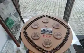 Okrągła, drewniana tablica pamiątkowa z motywami kolejowymi, eksponowana w przeszklonej gablocie.