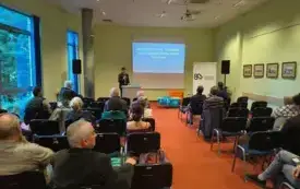 Na pierwszym planie są uczestnicy spotkania. Na drugim planie w centrum sw&oacute;j referat wygłasza Łukasz Zborowski. Stoi przy m&oacute;wnicy, po prawej stronie znajduje się stolik z promocyjnymi torbami EDD. W tle wyświetlona jest prezentacja.