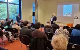 Na pierwszym planie znajdują się uczestnicy spotkania. Słuchają Dyrektora Biblioteki otwierającego spotkanie. Za nim wyświetlony został plakat wydarzenia, a po prawej stronie widać baner z logiem Biblioteki informujący także o jej 80-leciu.